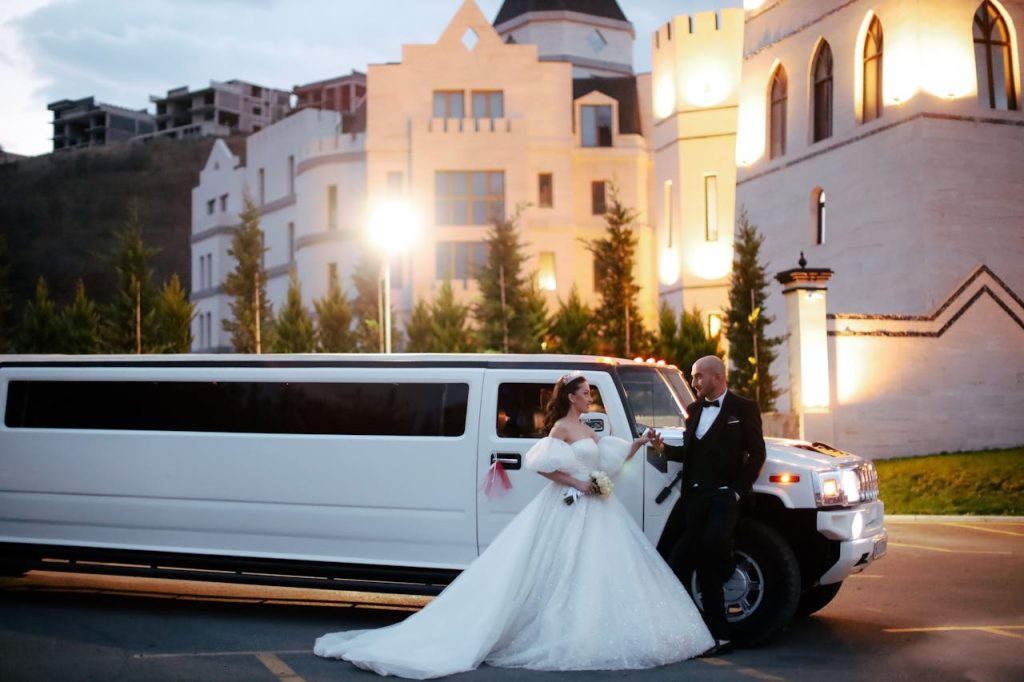 newlyweds-standing-together-by-limousine-20297470 Bride and groom in formal attire with a limousine in front of an illuminated building at night.
